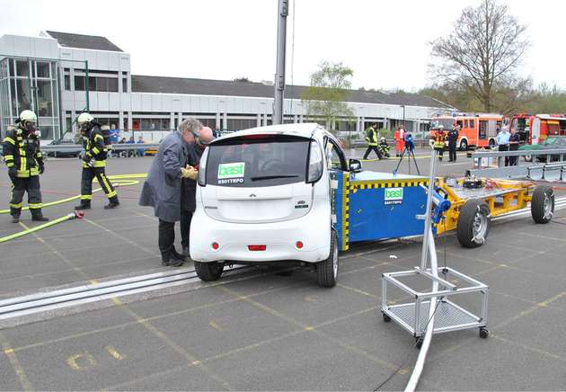 Nach dem Crash-Test eines Elektroautos wird der mögliche Austritt von gefährlichen Batteriegasen wie Fluorwasserstoff direkt im Batteriebehälter gemessen. Eine silberne Gasprobenahmeleitung führt zum Messgerät.