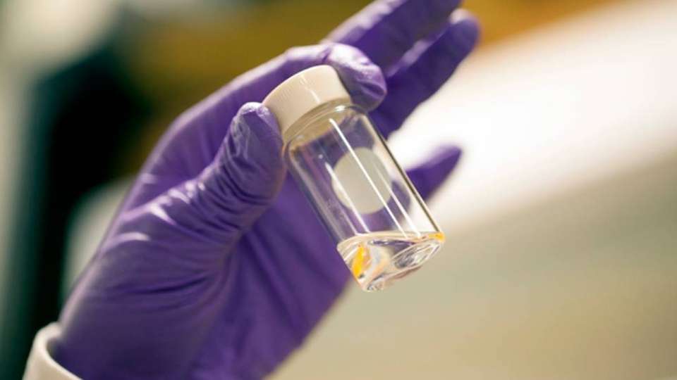Die Doktorandin Ingrid Guha hält ein Glas mit einer klaren Flüssigkeit, die mit bloßem Auge wie Wasser aussieht, aber  eigentlich eine Emulsion aus Öl und Wasser im Nanobereich ist.