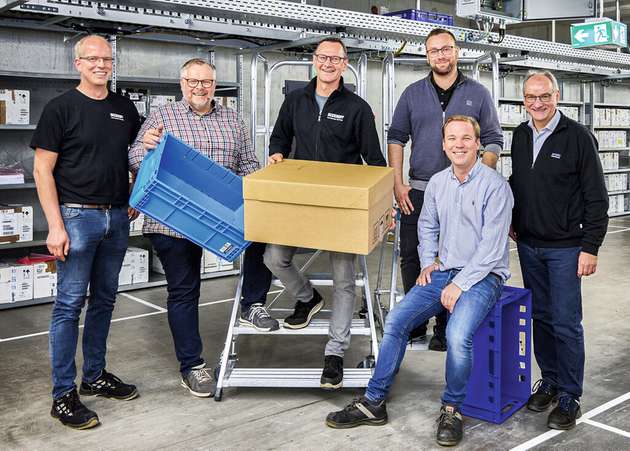 Stefan Maßmann, Jürgen Bolte und Dimitri Kool (alle Beckhoff Anlagentechnik), Maximilian Budde (sitzend) und Tristan Rodewald (beide Budde Systems) und Markus Wolharn (Arvato SE) – v.l.n.r. – vor dem unter der Decke montierten Ringförderer mit einem der 27 Schaltschränke zur Versorgung der EP7402.