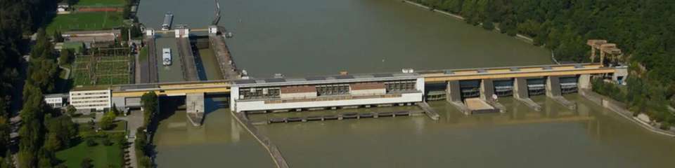 Luftaufnahme des Donaukraftwerks Aschach mit Wehranlage und Österreichs tiefster Donau-Schiffsschleuse, eingebettet in die enge Flusspassage zwischen bewaldeten Hängen.